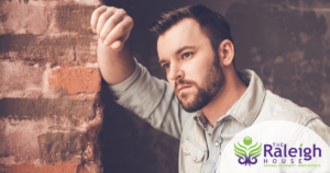 A bearded middle-aged man wearing a denim shirt rests his arm on a brick wall, appearing deep in thought.