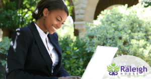 A woman sits in the sun outside, working on her laptop.