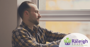 A middle-aged man sits near a window looking out with a forlorn look on his face.