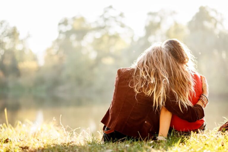 Two women sitting at a lakeshore in the sunshine.